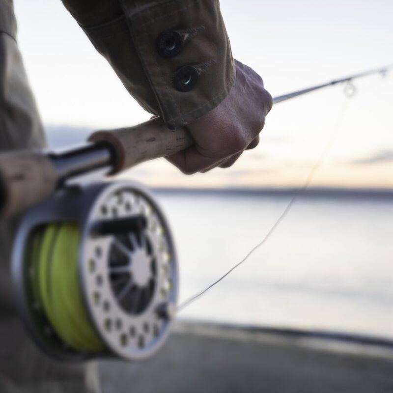 A closeup of a male hand holding a fly fishing rod and reel at the water's edge on a beach.