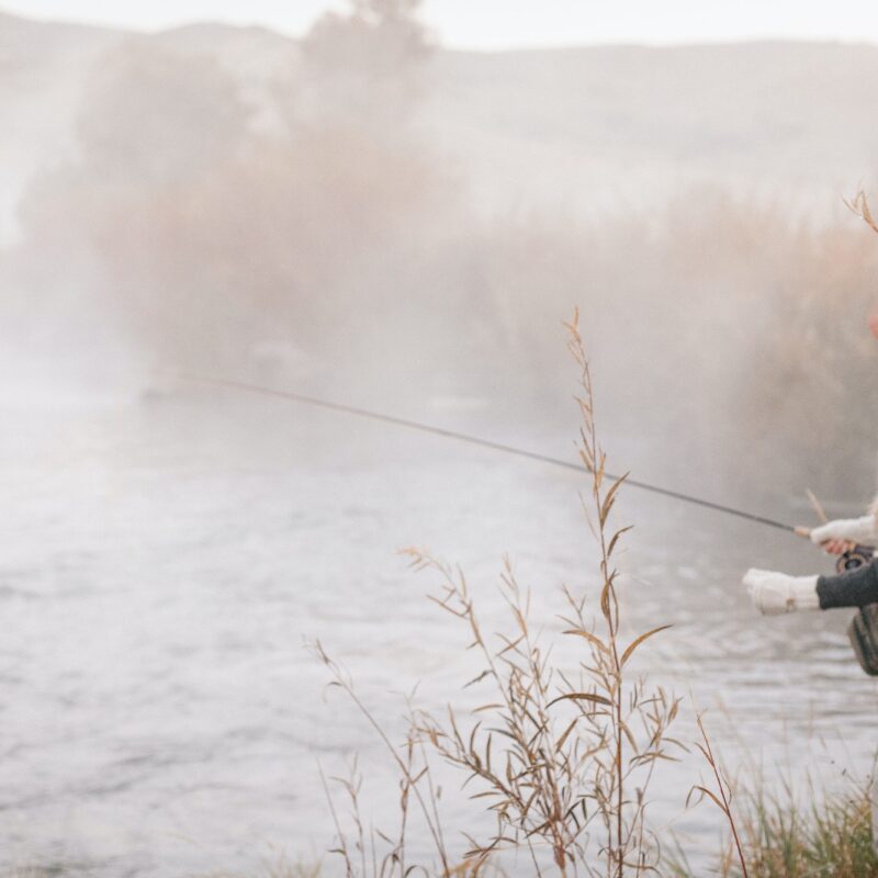 A fisherman, a woman standing on the banks of a river, flyfishing.