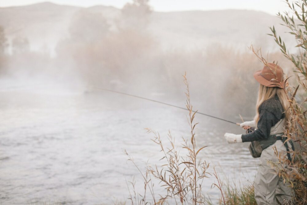 A fisherman, a woman standing on the banks of a river, flyfishing.