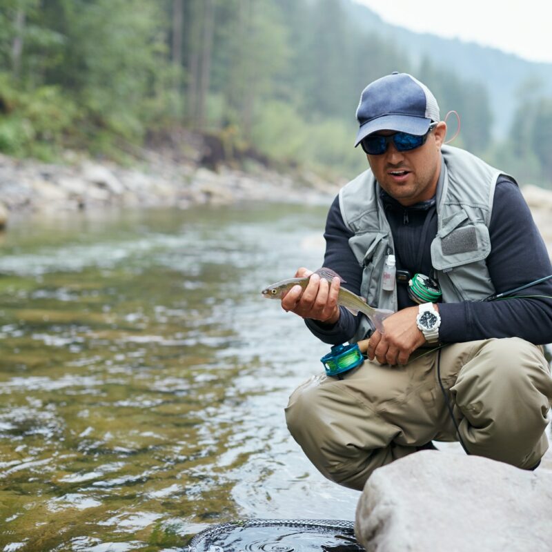 Happy fisherman holding trout while sitting near river