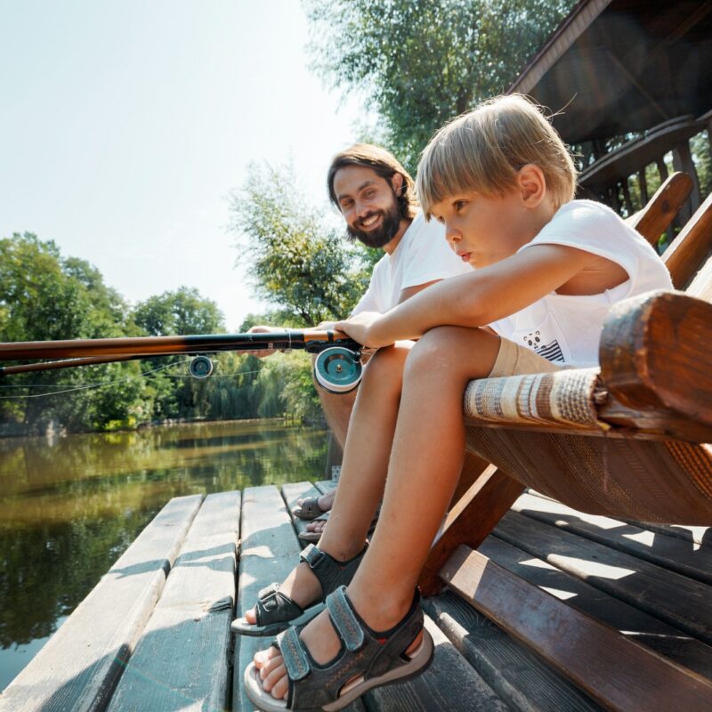 Little blond boy and his handsome father are sitting in recliners on the wooden pier and fishing