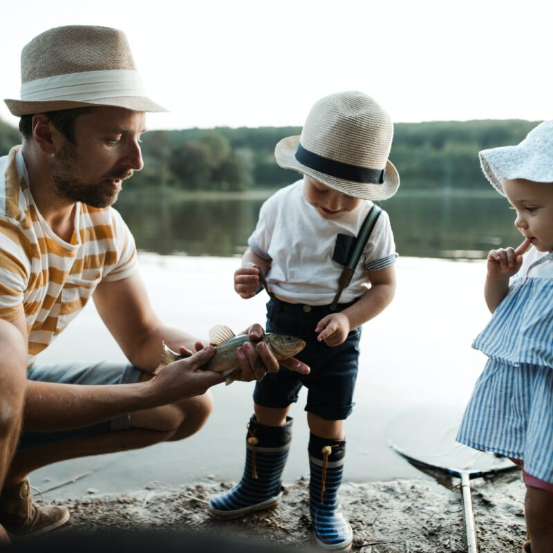 A mature father with small toddler children fishing by a lake, holding a fish.