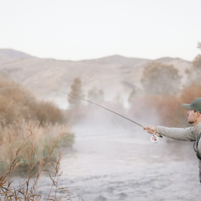 A man standing casting his fishing rod, flyfishing from a riverbank.