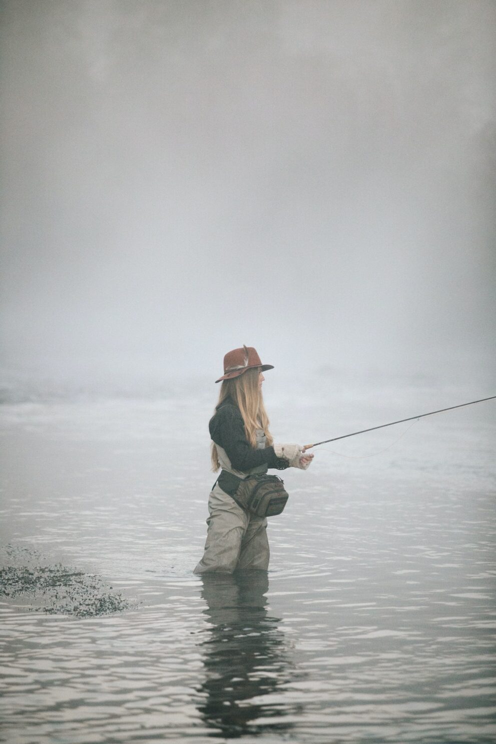 A woman fisherman flyfishing, standing in waders in thigh deep water.